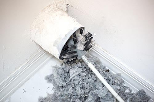 Dryer vent in a home being cleaned out with a round brush. There is a large pile of lint that has been removed from the vent on a white tiled floor. The walls and baseboards are white. The lint is gray. Taken with a Canon 5D Mark 3 camera. rm